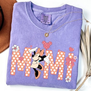 Könnte beinhalten: Ein lila Comfort Colors T-Shirt mit einem Minnie Mouse-Motiv. Das Wort "Mimi" ist in großen, gepunkteten Buchstaben gedruckt. Das Design umfasst Herzen, Sterne und florale Akzente. Eine goldene Halskette mit Anhänger liegt auf dem Shirt.
