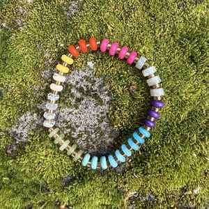 May include: A colorful beaded bracelet with a rainbow of hues, including red, orange, yellow, pink, blue, and purple. The bracelet features gold-colored spacers between the beads. The bracelet is displayed on a bed of green moss.
