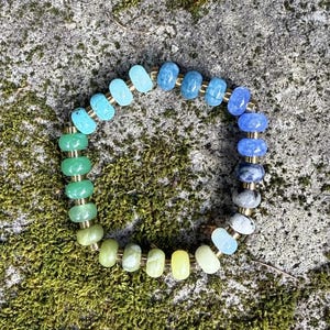 May include: A beaded bracelet with a gradient of colors, from green to blue, with gold-colored spacers. The bracelet is resting on a mossy, gray stone surface. The beads are round and smooth.