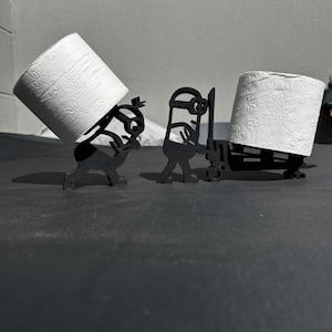 May include: Black metal toilet paper holders in the shape of Minions characters. One Minion is holding a roll of white toilet paper, and the other is pushing a pallet with a roll of toilet paper. The holders are on a dark surface.