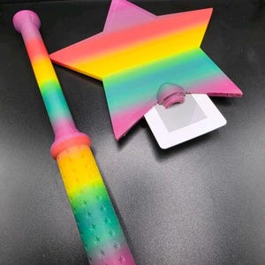 May include: A rainbow-coloured star-shaped object and a wand. The star has a slot for a card. The wand is cylindrical with a handle and a star pattern. The colours transition from pink to yellow to green.