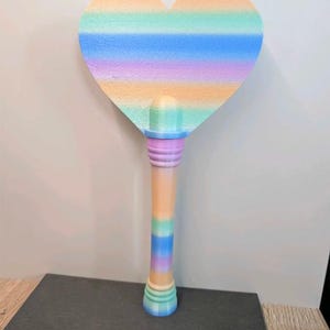 May include: A heart-shaped wand featuring a gradient of pastel colors including orange, yellow, green, blue, and purple. The handle is cylindrical with matching horizontal stripes in the same color palette, transitioning from a rounded top to a flared base.