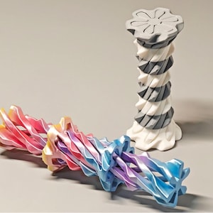 Può includere: Due sculture astratte. Una è una colonna alta e a spirale in bianco, grigio e con una parte superiore grigia. L'altra è una forma contorta multicolore con sfumature di rosa, giallo e blu. Le sculture sono su sfondo neutro.