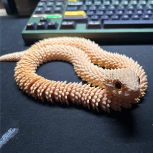 May include: A peach-coloured articulated snake toy with a textured, scale-like design. The snake's head has detailed eyes and a slightly open mouth. The toy is on a dark surface.