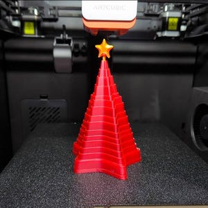 May include: A red, 3D-printed Christmas tree with a gold star on top. The tree is made of stacked layers and sits on a textured black surface. The Anycubic 3D printer is in the background.