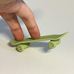 May include: A small, light green skateboard with four wheels. The skateboard is being held by a person's fingers. The skateboard is a miniature version of a full-sized skateboard.