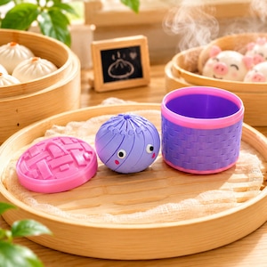 May include: A purple plastic dumpling-shaped container with a pink lid and a matching purple basket-weave base. The container features two small white eyes and pink cheeks. It sits on a round wooden tray alongside a separate pink lid.