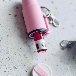 K&ouml;nnte beinhalten: Ein rosa Lippenstifthalter mit schwarzem Rand, der einen roten Lippenstift mit Kirschmuster enth&auml;lt. Der Halter ist ge&ouml;ffnet und zeigt den Lippenstift. Ein wei&szlig;er Deckel mit rosa Riemen ist in der N&auml;he. Ein Schl&uuml;sselanh&auml;nger ist ebenfalls sichtbar.