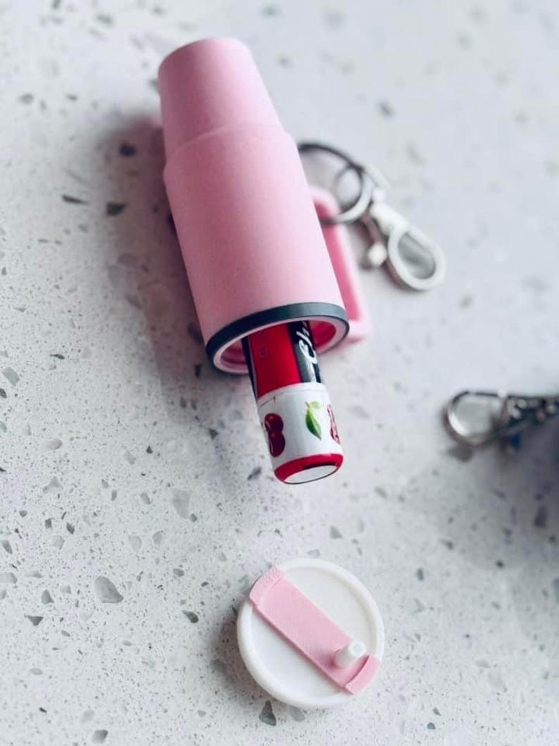 K&ouml;nnte beinhalten: Ein rosa Lippenstifthalter mit schwarzem Rand, der einen roten Lippenstift mit Kirschmuster enth&auml;lt. Der Halter ist ge&ouml;ffnet und zeigt den Lippenstift. Ein wei&szlig;er Deckel mit rosa Riemen ist in der N&auml;he. Ein Schl&uuml;sselanh&auml;nger ist ebenfalls sichtbar.