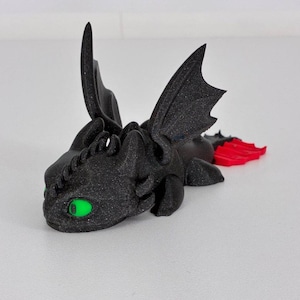 Puede incluir: Figura de dragón negro con ojos verdes y detalles rojos en la cola. El dragón tiene alas detalladas y una superficie texturizada, lo que le da una apariencia impresa en 3D. La figura está tumbada sobre una superficie blanca.