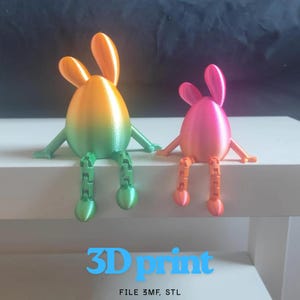 May include: Two 3D-printed bunny figures. One is a gradient of green and orange, the other is pink and orange. Both have articulated legs and arms, and are sitting on a white surface. The text "3D print" and "FILE 3MF, STL" are at the bottom.