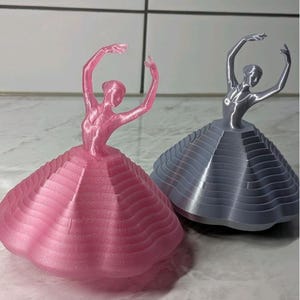 Pode incluir: Duas estatuetas de bailarina. Uma é rosa, a outra cinza. Ambas têm os braços levantados acima da cabeça. As saias são em camadas e em forma de cone. As estatuetas estão sobre uma superfície clara.