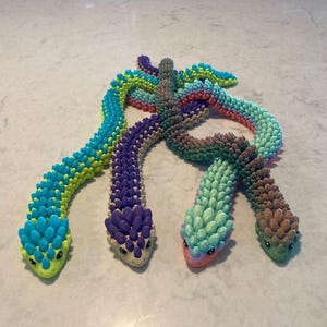 May include: Four colorful, handmade snake toys in shades of blue, green, purple, and brown. Each snake has a detailed, textured body and a small, detailed head. The snakes are arranged in a playful, intertwined pattern.