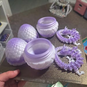 May include: A collection of 3D-printed items in shades of purple and white. The items include two round containers, two dragon-scale egg-shaped objects, and two dragon-shaped ornaments. The objects are displayed on a brown surface.