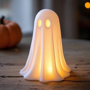 Puede incluir: Lámpara blanca con forma de fantasma con dos aberturas ovaladas para los ojos. La lámpara emite un brillo amarillo cálido. Una calabaza naranja es visible en el fondo. Decoración temática de Halloween.