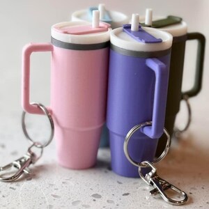 K&ouml;nnte beinhalten: Miniatur-Becher-Schl&uuml;sselanh&auml;nger in Rosa, Lila, Blau und Olivgr&uuml;n. Jeder Schl&uuml;sselanh&auml;nger hat einen kleinen Griff, einen wei&szlig;en Deckel mit Strohhalm und einen silbernen Verschluss. Die Schl&uuml;sselanh&auml;nger sind so gestaltet, dass sie Reisebechern &auml;hneln.