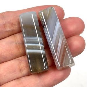May include: Two rectangular, polished agate stones with a mix of gray, white, and brown bands. The stones are held in a hand, showcasing their smooth, glossy surfaces. These stones are ideal for jewelry making or collecting.