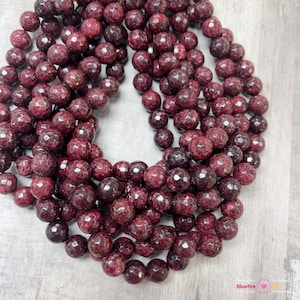 May include: A strand of dark red faceted gemstone beads. The beads are round and have a rough, textured surface.