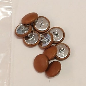May include: A collection of tan-colored buttons with silver metal backs. The buttons are in a pile next to a clear plastic bag with a handwritten label that reads "30L(3/4) 4PL TAN 10".