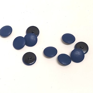 Puede incluir: Diez botones redondos de color azul oscuro con centros negros. Los botones están dispersos sobre una superficie blanca.