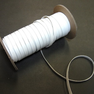 May include: A spool of white elastic ribbon. The ribbon is tightly wound around a cardboard core, with a length of the ribbon extending out. The elastic is a bright white color and appears smooth.
