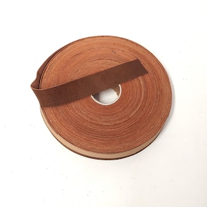 Może przedstawiać: Rolka brązowej wstążki z ciemniejszym brązowym paskiem na wierzchu. Wstążka jest ciasno zwinięta, ukazując krawędzie każdej warstwy. Tło jest gładko białe.