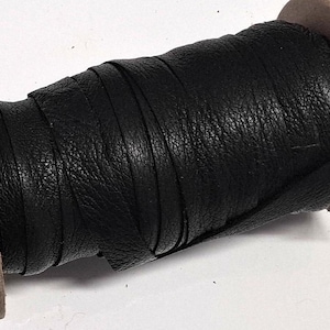 May include: A spool of black leather cord wrapped around a cylindrical cardboard core. The cord has a textured surface and is wound tightly in multiple layers across the length of the spool, showing a consistent matte black finish throughout.