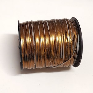 Może przedstawiać: Szpula płaskiego skórzanego sznurka w metalicznym kolorze miedzi. Sznurek jest ciasno nawinięty na czarną plastikową szpulę. Skóra wydaje się cienka i elastyczna, z błyszczącą, odblaskową powierzchnią. Szpula jest na białym tle.