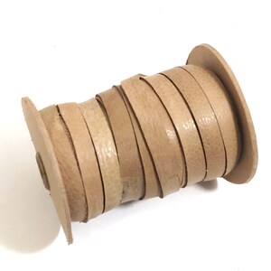 Op de afbeelding: Een spoel met lichtbruin lederen koord. Het koord is strak om de spoel gewikkeld en klaar voor knutselprojecten.