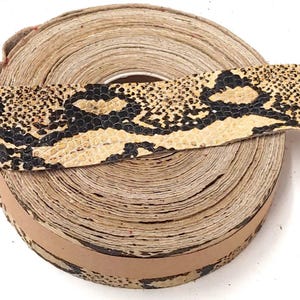 May include: A coiled strip of leather trim featuring a textured snakeskin pattern in shades of cream, beige, and black. The material is wound into a tight circular roll, displaying the repeating reptilian scale print across the outer surface.