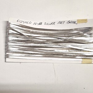 Może przedstawiać: Szpula srebrnej metalicznej wstążki, ciasno nawinięta i przymocowana do białej karty. Karta zawiera odręczny tekst: "0125NCD 10 YDS SILVER MET BRK". Wstążka ma jasny, błyszczący srebrny kolor.