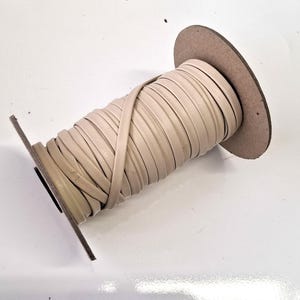 May include: A spool of beige leather trim. The trim is wound around a cardboard core, with the edges of the trim visible. The spool is set against a white background. The trim appears to be flat and flexible, suitable for various crafting projects.