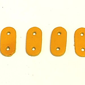 Puede incluir: Cuatro correas de cuero naranja con dos agujeros cada una. Las correas son de forma ovalada y tienen una textura lisa.