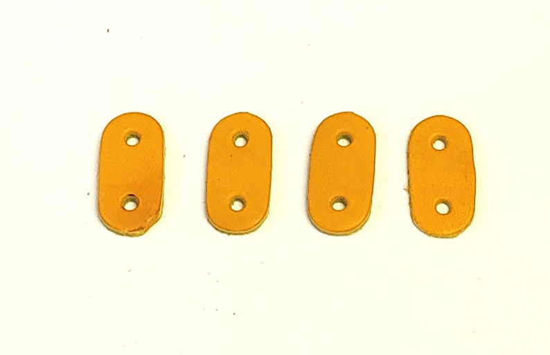 Puede incluir: Cuatro correas de cuero naranja con dos agujeros cada una. Las correas son de forma ovalada y tienen una textura lisa.