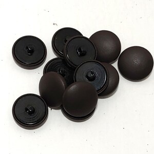 May include: A collection of dark brown, round buttons with a matte finish. Each button has a black, recessed back with a loop for sewing. The buttons are scattered on a white surface, showcasing their simple design.