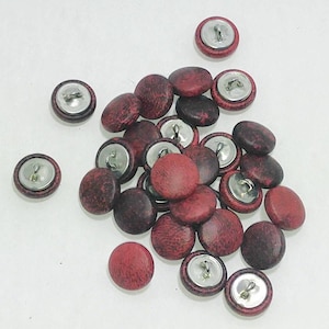 May include: A collection of round buttons featuring a mottled red and black surface pattern. Each button has a smooth, circular top and a silver-toned metal shank on the underside for attachment. The buttons are scattered in a loose cluster.