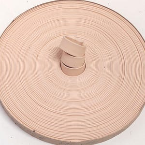 May include: A coiled roll of light beige vegetable-tanned leather strip. The material is smooth and uniform in color, wound tightly into a circular shape with a small section of the strip protruding from the center of the coil.