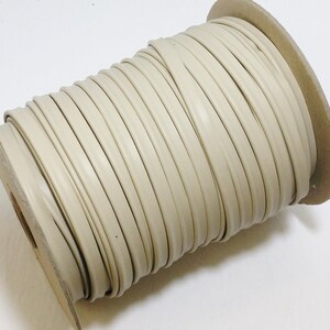 May include: A spool of beige piping cord wrapped around a cardboard cylinder. The cord is smooth, rounded, and features a consistent light tan color throughout the entire length of the material wound tightly onto the central core.