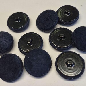 May include: A collection of ten round buttons featuring a dark navy blue fabric-covered top surface and a smooth black plastic base with a central shank attachment point on the back, arranged in a scattered pattern on a neutral surface.