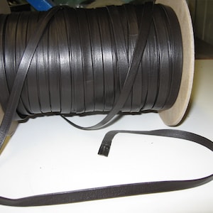 May include: A spool of brown leather cord, approximately 1/4 inch wide, is wound around a cardboard core. The cord is unraveled and laid out on a white surface.