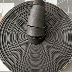 以下が含まれることがあります： 黒いゴム紐のロールで、同心円状にきつく巻かれています。同じ紐のカットされた部分のスタックがロールの上にあり、質感と厚みを示しています。この紐は、耐久性があり柔軟な素材で作られているようです。