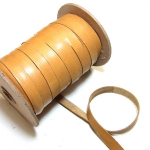 Puede incluir: Un carrete de cordón de cuero de color marrón claro, enrollado alrededor de un carrete de madera. El cordón tiene aproximadamente 6 mm de ancho y se muestra desenrollado en un bucle.