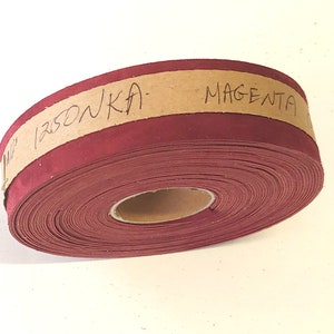 Op de afbeelding: Een rol bordeauxrode stofband met een bruin papieren label met de tekst "1250NKA - MAGENTA".