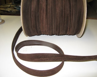 May include: A spool of dark brown suede trim. The trim is folded and rolled onto a cardboard spool. A length of the trim is unwound and lies in a loop in the foreground. The trim is a solid, rich brown color.