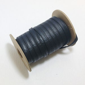 Puede incluir: Un carrete de cordón elástico negro enrollado alrededor de un carrete de cartón. El cordón es texturizado y tiene un ligero brillo.