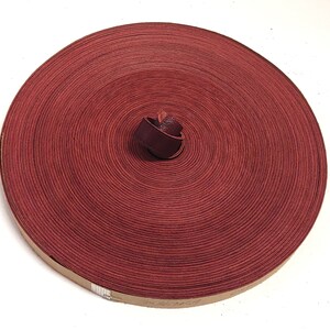 Op de afbeelding: Een rol rood lederen koord, opgerold tot een cirkel. Het koord is ongeveer 1,2 cm breed en heeft een gladde, gepolijste afwerking.