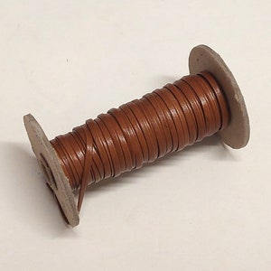 May include: A spool of brown leather cord wrapped around a cardboard core. The cord is approximately 1/4 inch wide.