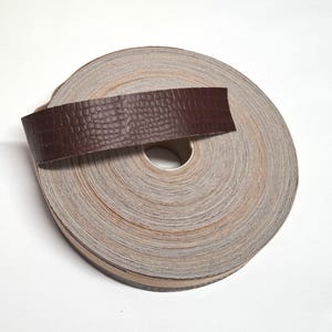 May include: A roll of brown leather trim with a crocodile pattern. The trim is wrapped around a spool of beige fabric.