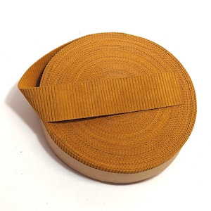 Może przedstawiać: Rolka ozdobnej taśmy z żebrowanej tkaniny w kolorze musztardowym. Taśma jest zwinięta i związana kawałkiem tej samej tkaniny.