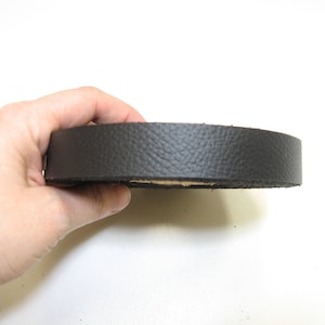 Może przedstawiać: Zbliżenie czarnego skórzanego paska, zwiniętego i trzymanego w dłoni. Pasek jest gładki i ma lekko fakturowaną powierzchnię.